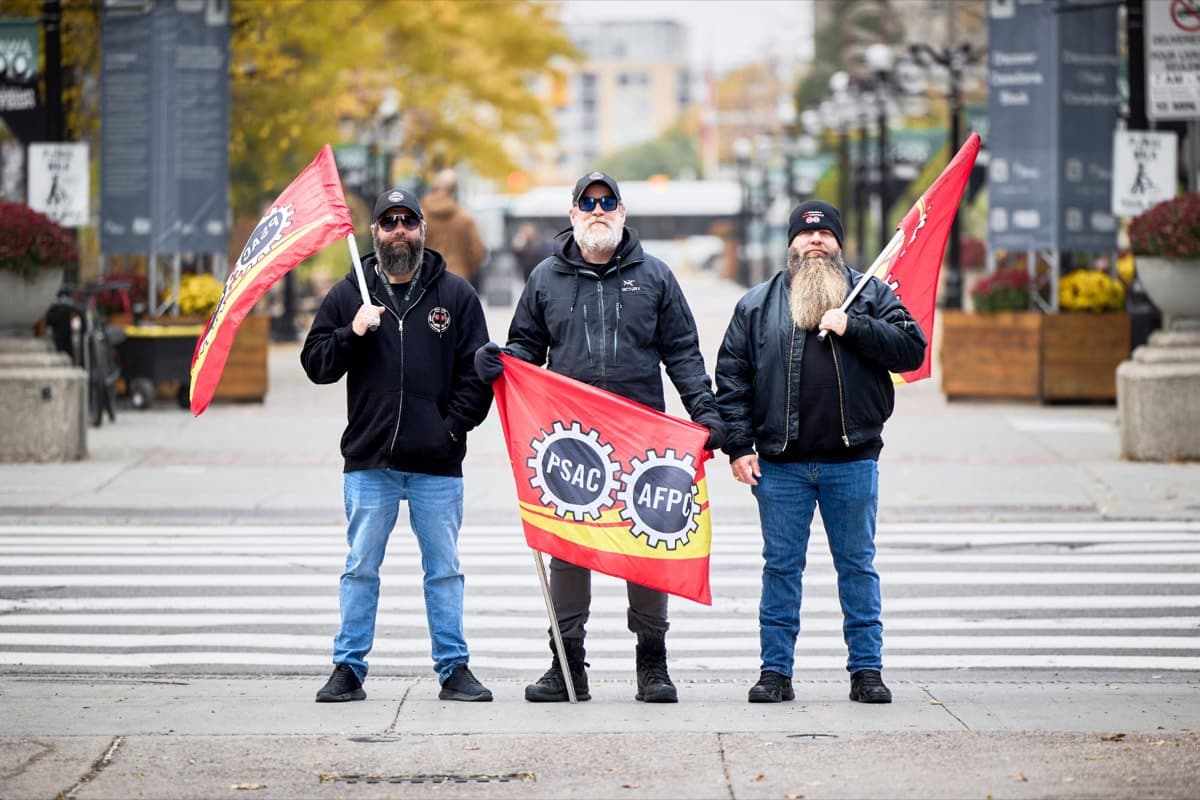 Trois piqueteurs se tiennent sur un passage pour piétons en tenant des drapeaux de l'AFPC-PSAC.